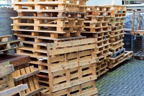 A large collection of wooden pallets stacked neatly outdoors on a paved surface, with some pallets leaning against others. The pallets are light-colored, showing natural wood grain and textures, with visible slats and support beams. In the background, a chain-link fence and a brick wall suggest an industrial or storage area, likely part of a waste management or rubbish removal site serving Sidcup or nearby areas. The pallets are arranged in multiple tall columns, some with slightly weathered finishes, and the scene is well-lit with natural daylight, emphasizing the structural details and textures of the wood. This arrangement of pallets reflects the type of rubbish material handled by Garden Clearance Sidcup when managing waste and rubbish removal for commercial or garden clearance services in the local district.