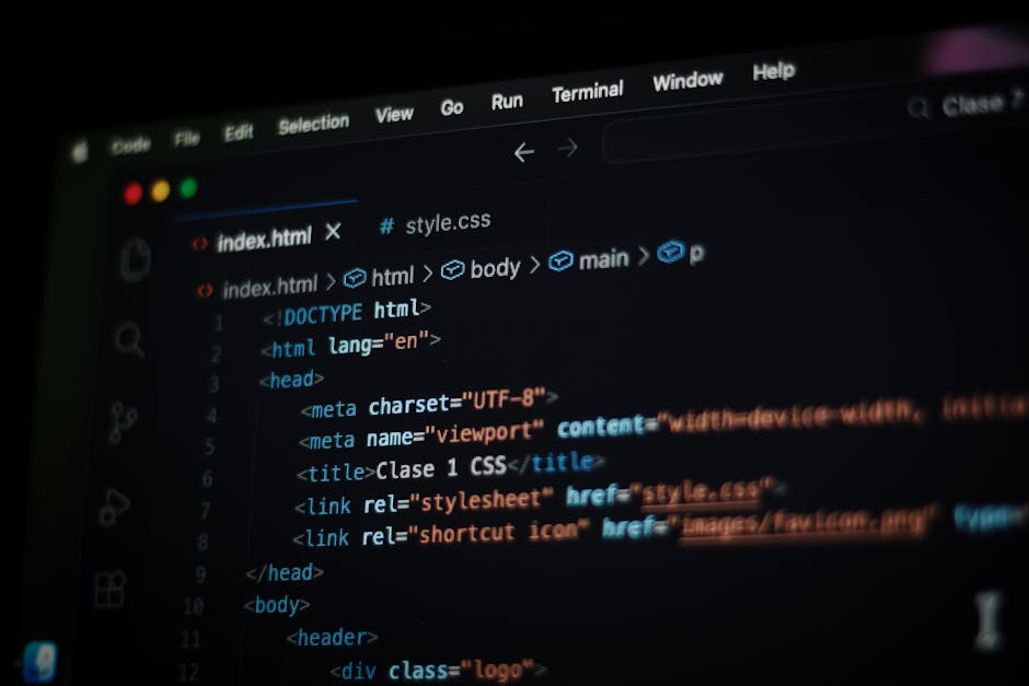 A young man with dark, curly hair and light skin is seated at a desk in a modern office environment, working intently on a computer with a large monitor displaying lines of code. The desk surface is c